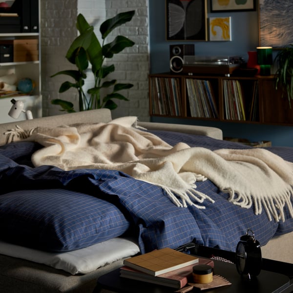 Lit douillet avec linge de lit à carreaux bleus, plaid blanc et plante verte en pot dans une chambre à coucher lumineuse.