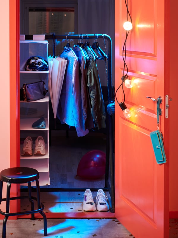 Un couloir avec un portant NIKKEBY où sont suspendus ou posés des vêtements, des chaussures et une planche à roulettes. Divers objets sont pendus à des crochets à proximité.