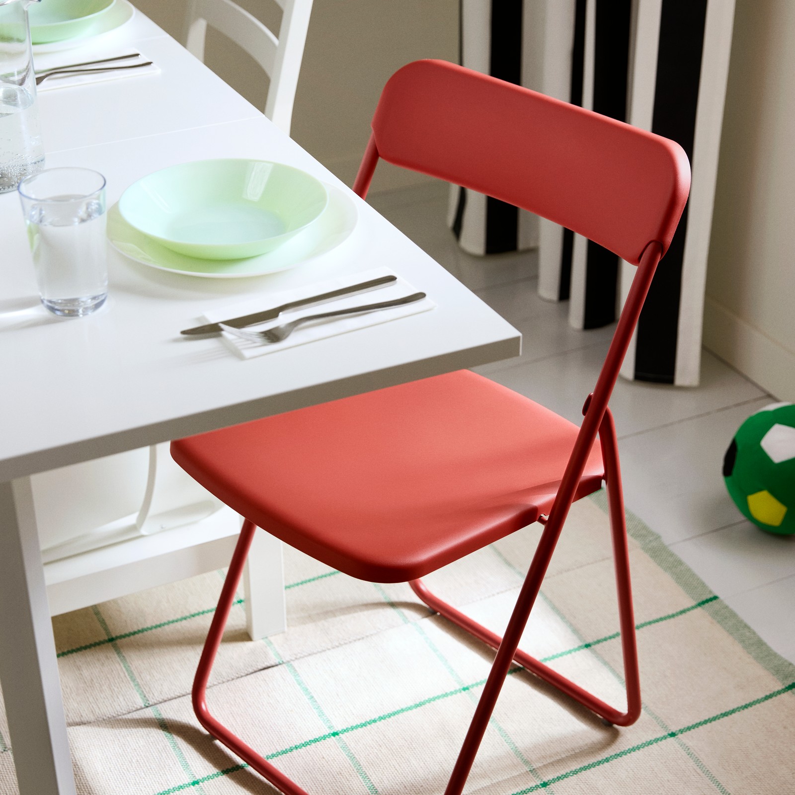 A foldable VIHALS chair in red, standing beside a white table that is set with cutlery, a bowl on a plate and a glass.