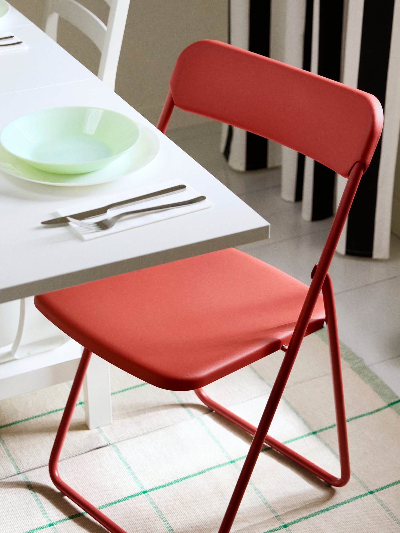 A foldable VIHALS chair in red, standing beside a white table that is set with cutlery, a bowl on a plate and a glass.