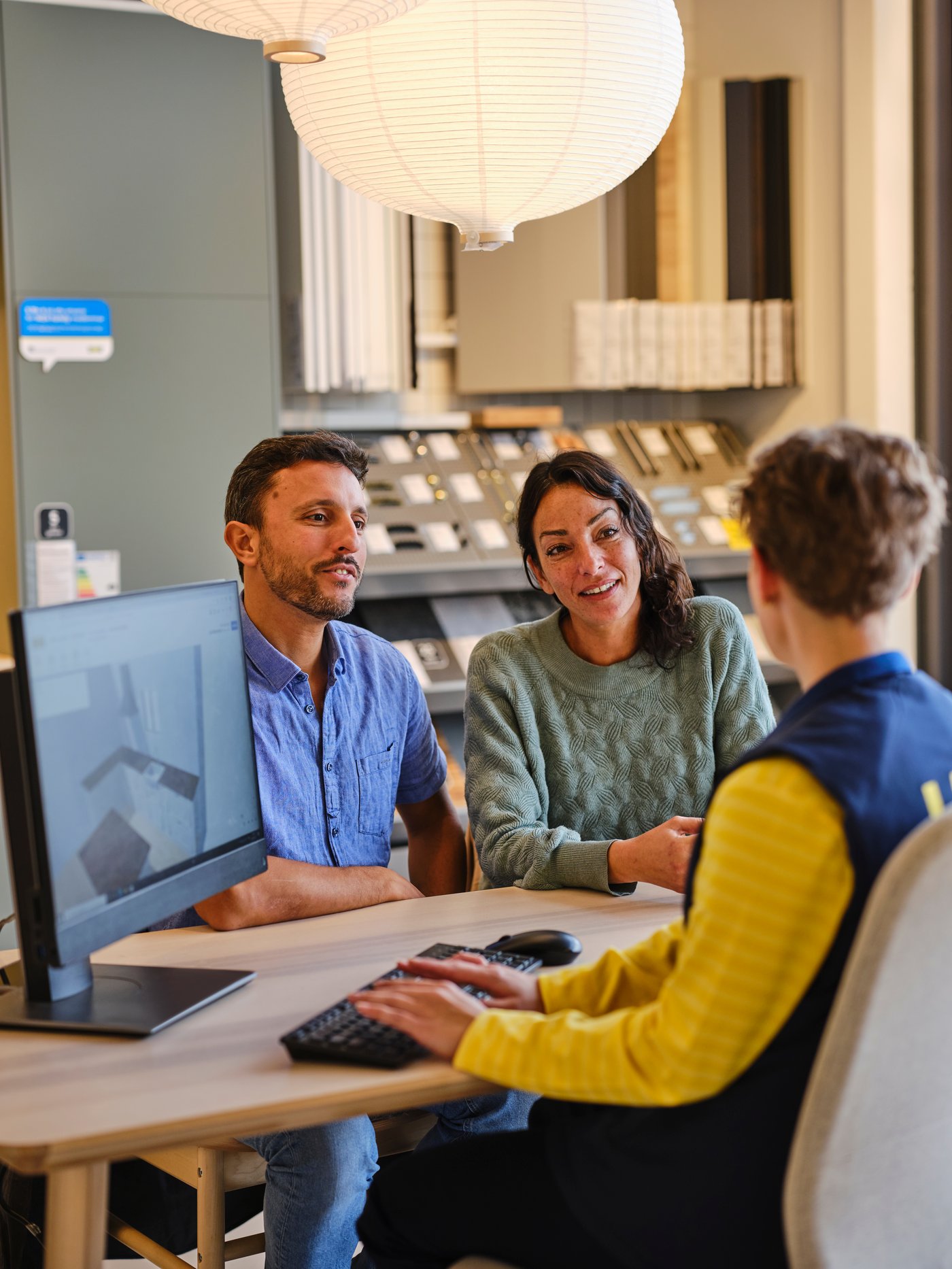 An IKEA co-worker wearing a uniform is talking with two customers in front of a computer.