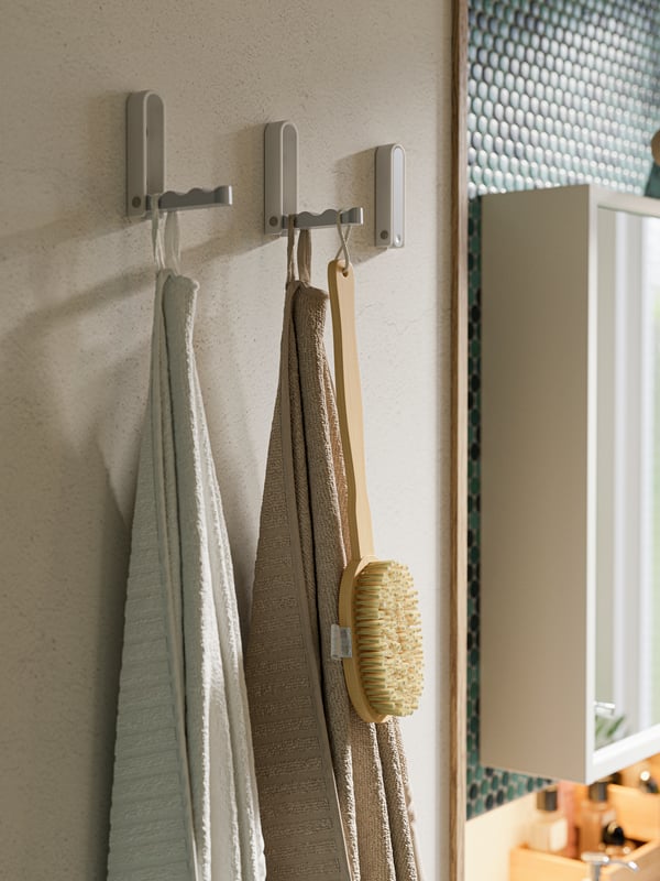 Tre ganci pieghevoli alla parete, un asciugamano beige chiaro, un asciugamano bianco e una spazzola da doccia in legno - IKEA.