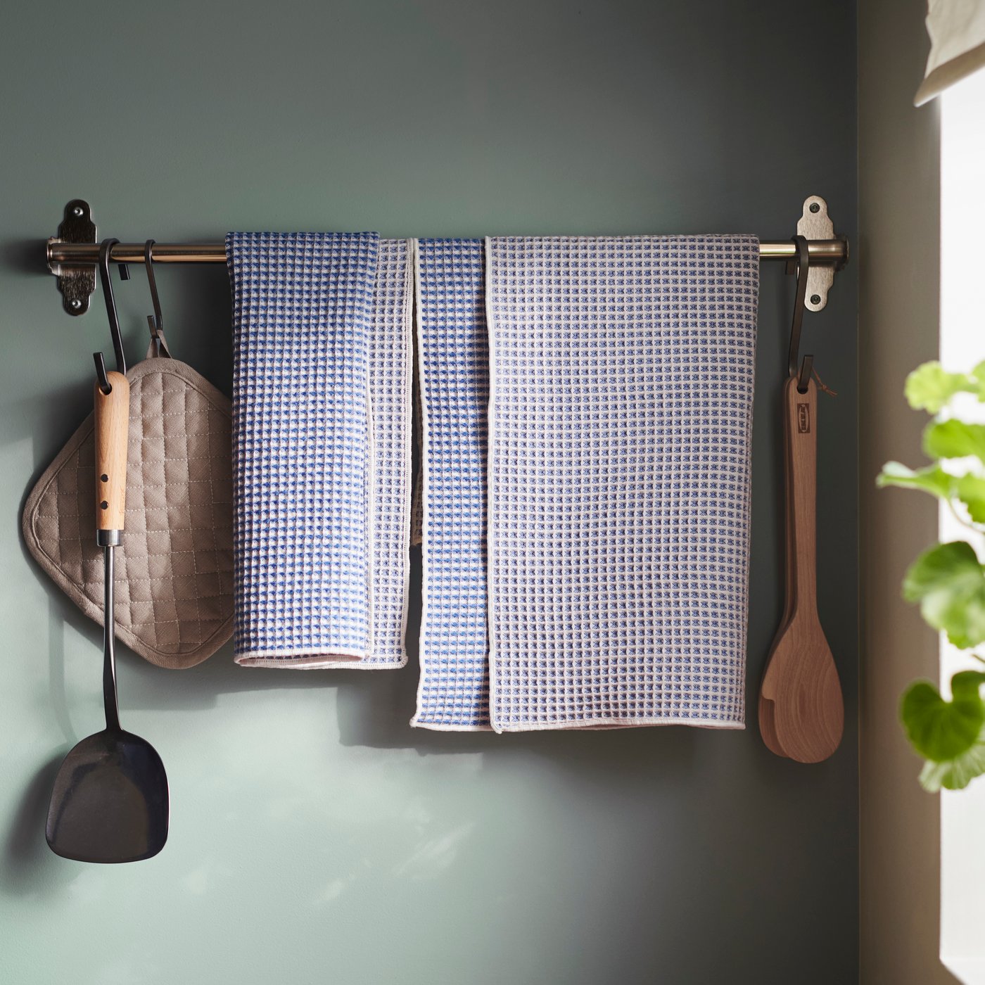 På en stång mot en grön vägg hänger olika kökshanddukar och köksredskap.