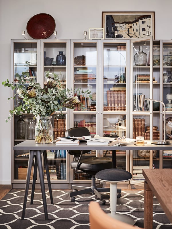 A NÄRSPEL desk with flowers in a vase and documents on top and three BILLY bookcases behind filled with books and mementos.