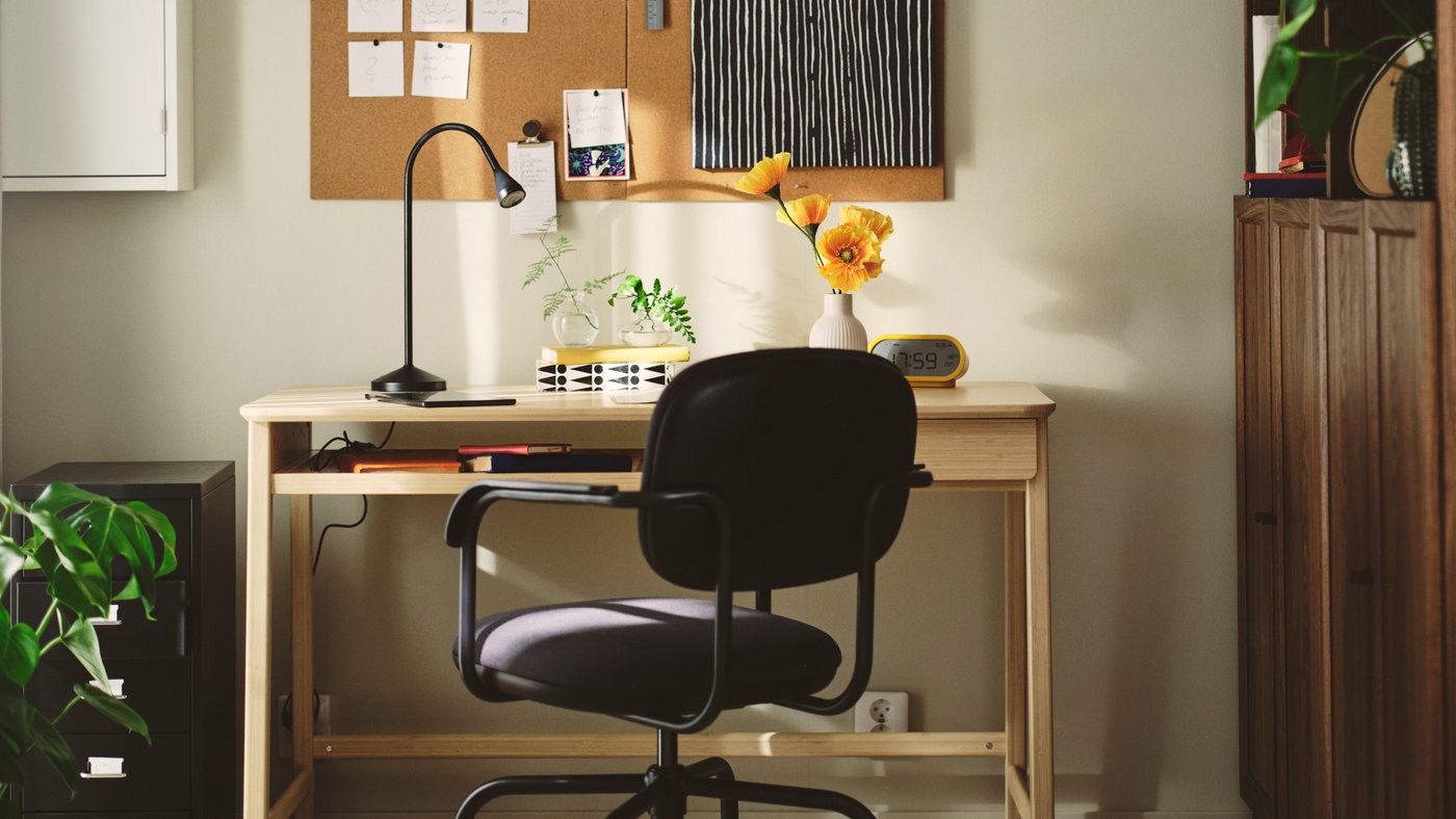 A home office with a dark grey MULLSJÖ swivel chair beside a light wooden desk and a cork board on the wall.