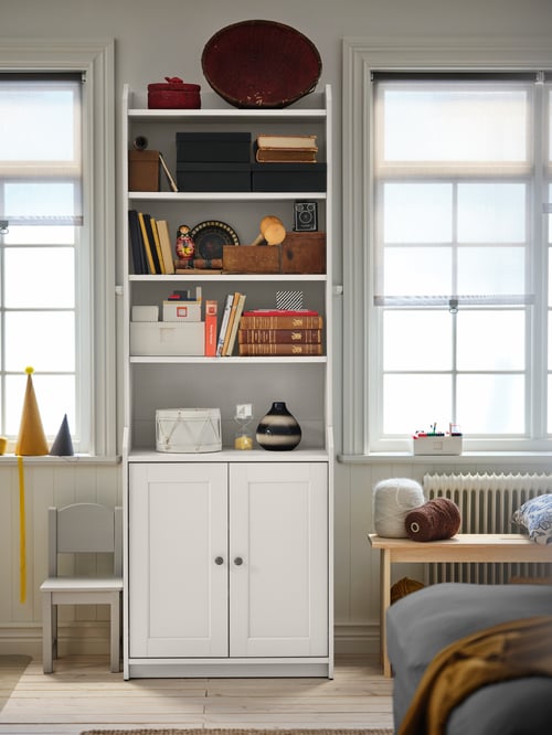 Une armoire haute blanche à deux portes contenant des livres et un ensemble de boîtes LEGO, une chaise pour enfants et un banc sont placés dans un salon.