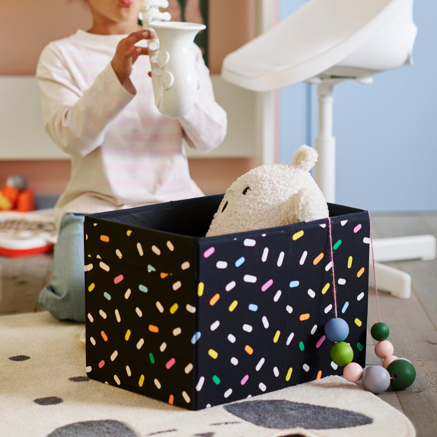 A black SKÅJARE box with colourful pattern is on the floor, with a white plush toy inside and a child playing nearby.