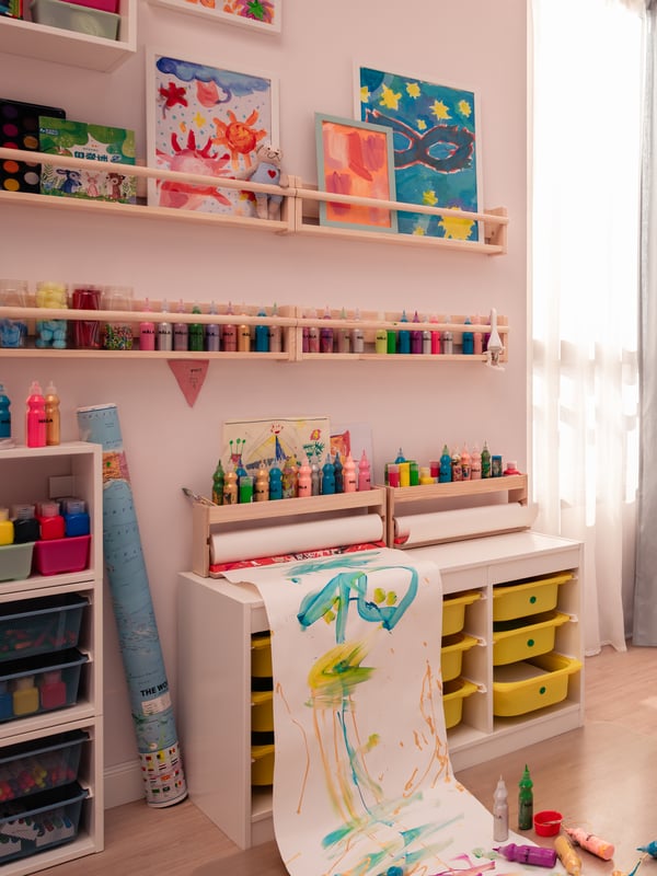 The image displays a well-organized children’s art corner. On the left, there is a storage unit with multiple transparent drawers filled with various art supplies. Next to it, a small white desk with shelves above holds numerous paint bottles neatly arranged by color. Above the desk, two shelves showcase colorful children’s paintings. A white chair with green and blue paint smears is in front of the desk, suggesting recent use for an art project. The room has a warm and creative atmosphere, inviting engagement in artistic activities.
