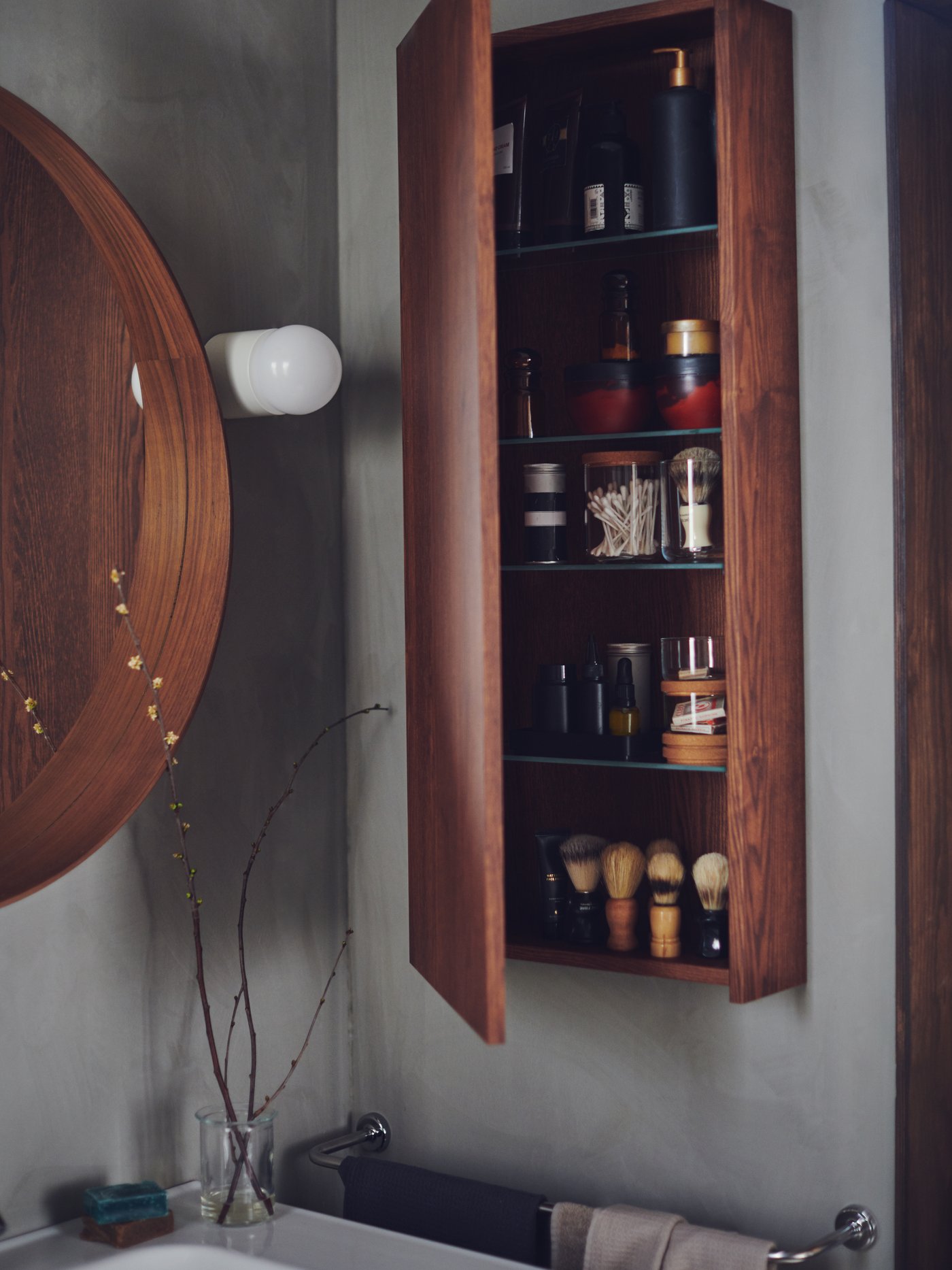 Smeđi GODMORGON ormarić na zidu kupatila iza čijih se otvorenih vrata vide police s toaletnim priborom i dodacima za brijanje.