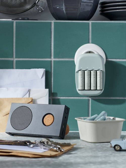Eine Arbeitsplatte mit einem eingesteckten STENKOL Ladegerät, weiteren Batterien in einer NOJIG Sortierbox darunter und einem ENEBY Bluetooth-Lautsprecher an der Seite.