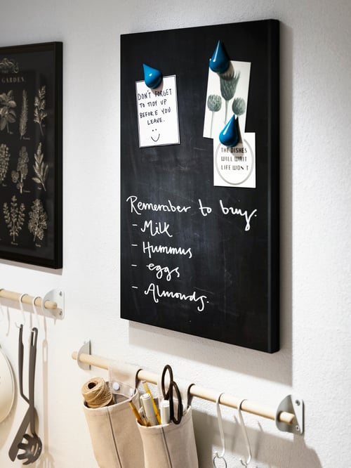 A section of a wall with a black SVENSÅS memo board with writing and notes. Below it hang accessories on a wall-mounted rail.