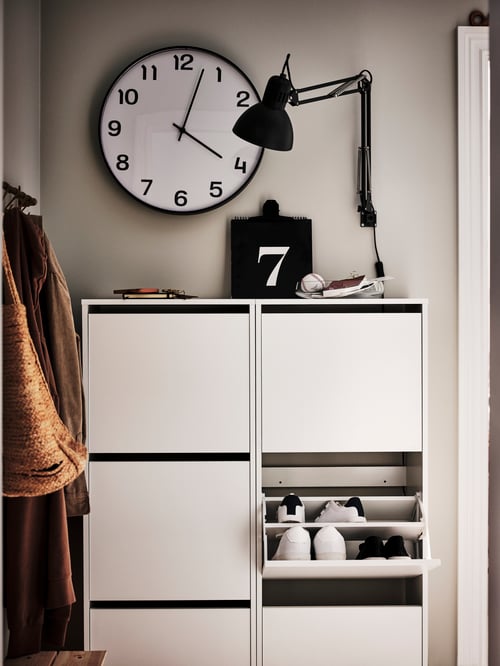 Hallway area featuring a two, white shoe cabinets with three compartments each, a wall clock, and a work lamp.