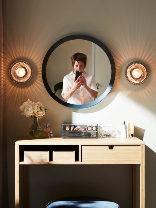 Bedroom with a person standing in front of a rectangular dressing table made of bamboo, storing 2 trays under a round mirror.