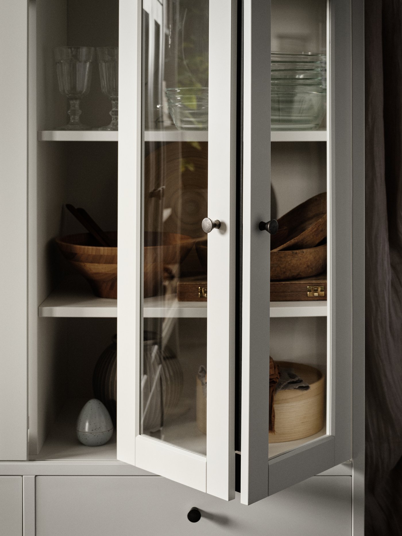 Un mueble blanco IDANÄS con puertas de vidrio plegadas, cristalería, vajilla de madera y una caja de chapa de fresno con tapa.