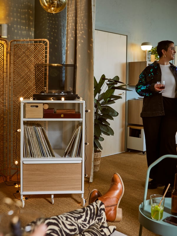 A softly lit space with people in relaxed poses in proximity to a white BEKANT storage unit holding LPs and a record player.