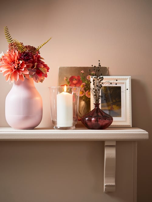 Une étagère avec une bougie blanche dans un vase en verre POMP, des fleurs dans des vases et des photos dans des cadres adossés au mur.