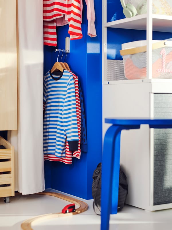 A KLYKET aluminium/beige folding hook on a blue wall near a white PLATSA open wardrobe, holding three t-shirts on hangers.