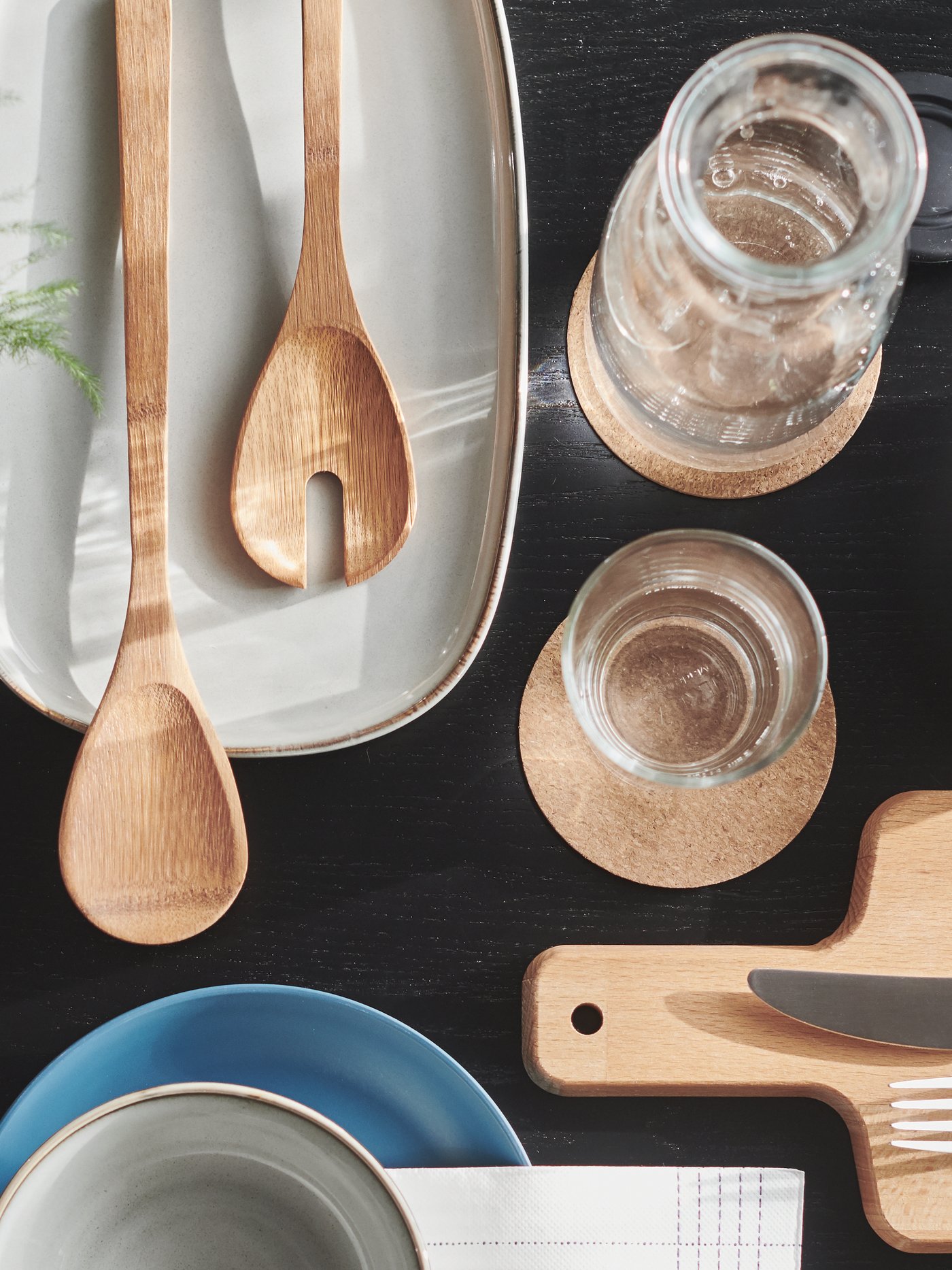 Vista superior de una mesa de comedor con un plato de gres gris, utensilios de madera, vaso y jarra sobre posavasos beige, juego de platos.