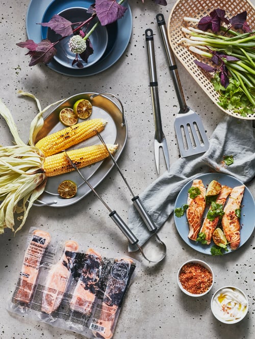 An unopened package of ASC-certified, frozen SJÖRAPPORT salmon fillets sits on a table by grilling supplies and vegetables.