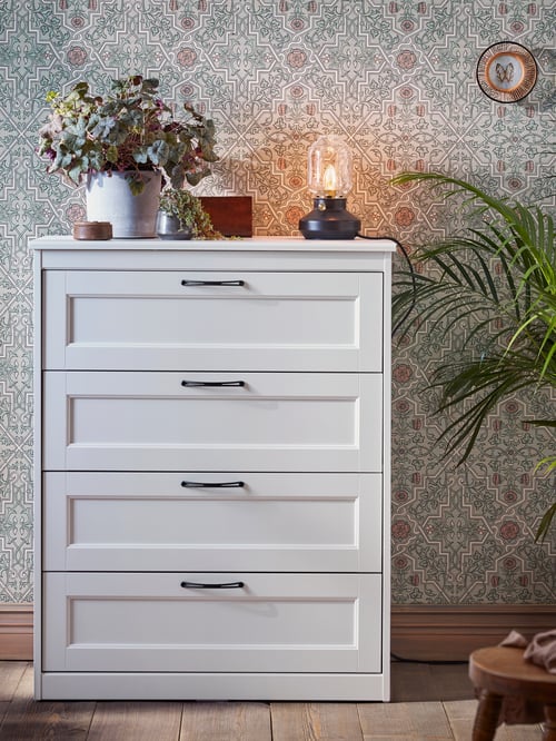Commode à quatre tiroirs SONGESAND blanche surmontée d’une lampe de table noire et installée devant un mur avec du papier peint à motifs.