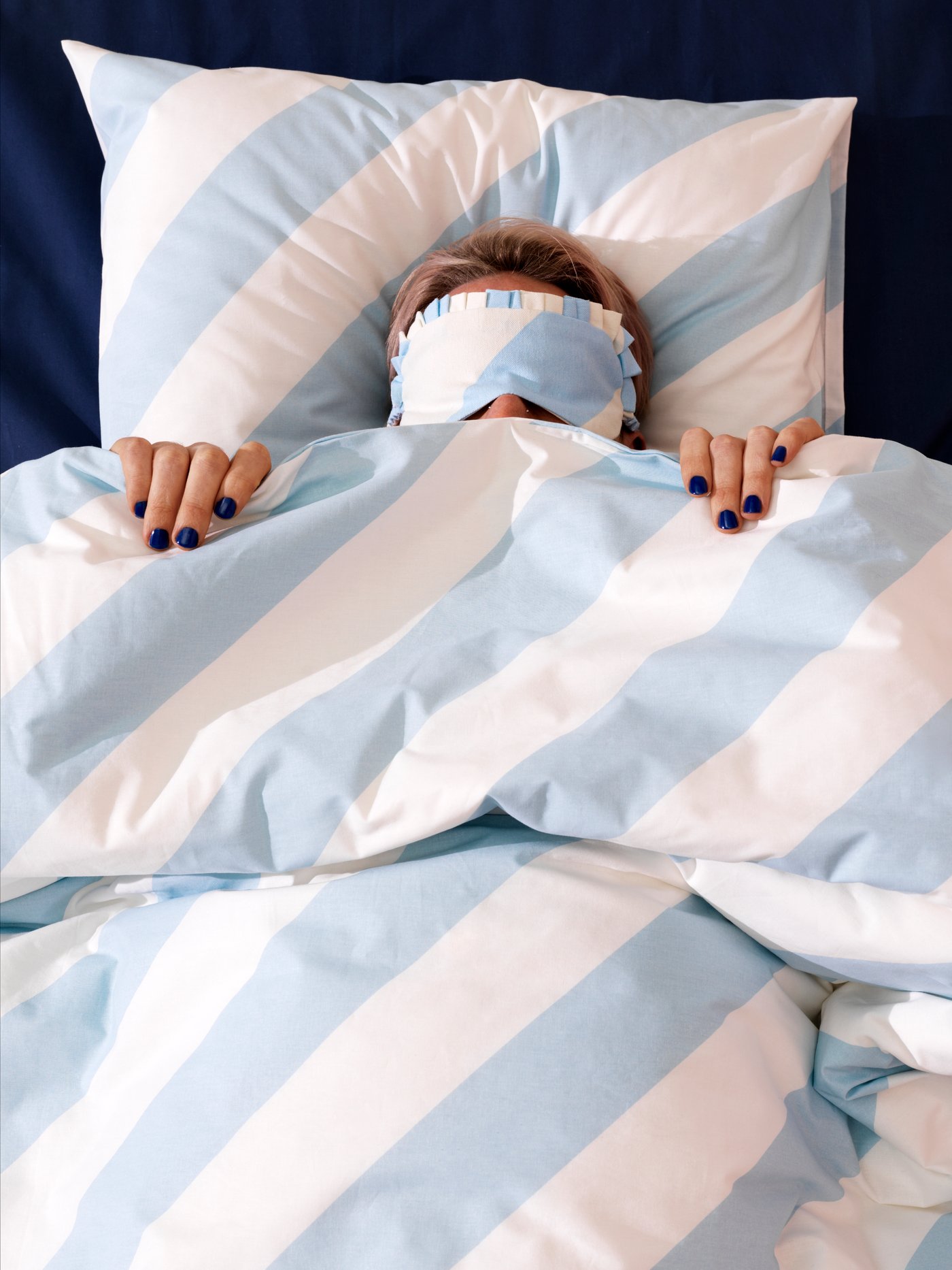 a person with an eye mask lying in a bed pulling a duvet over their face