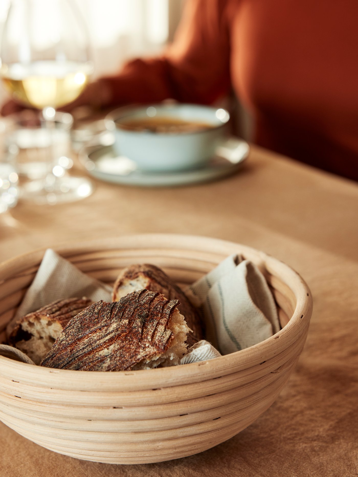 A person sits at a table which has a JÄSNING proofing/bread basket holding bread and some tableware on it.