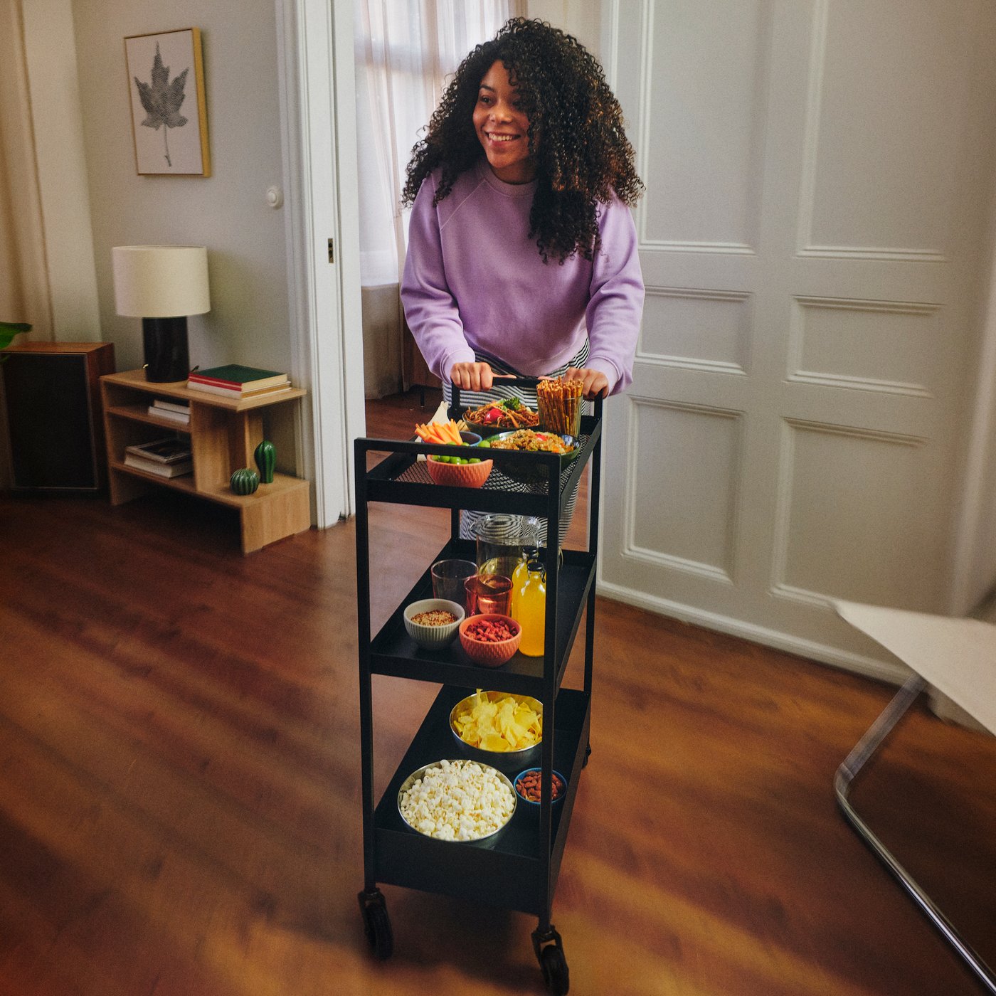 A person pushes a black NISSAFORS trolley with three metal mesh shelves. The trolley holds bowls of snacks and drink carafes.