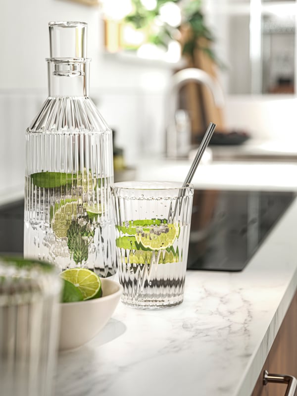 A bottle and a glass filled with water and lime wedges on an EKBACKEN worktop with a white lively marble effect/laminate.