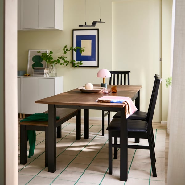 A bright dining area has an ÅLHULT extendable table in black/brown, with a matching bench and three chairs around it.