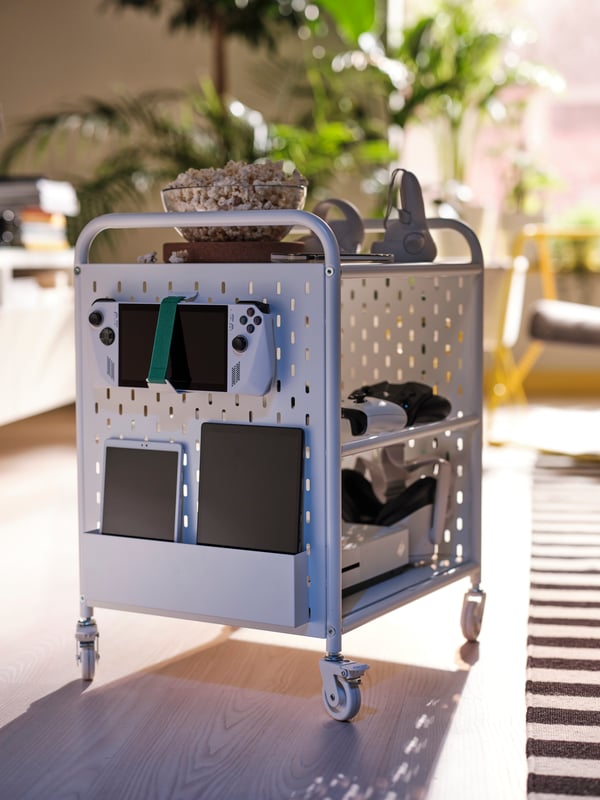 A white BRÄNNBOLL shelving unit on castors holds a popcorn bowl and gaming gear. More tech is stored on the unit’s pegboard.