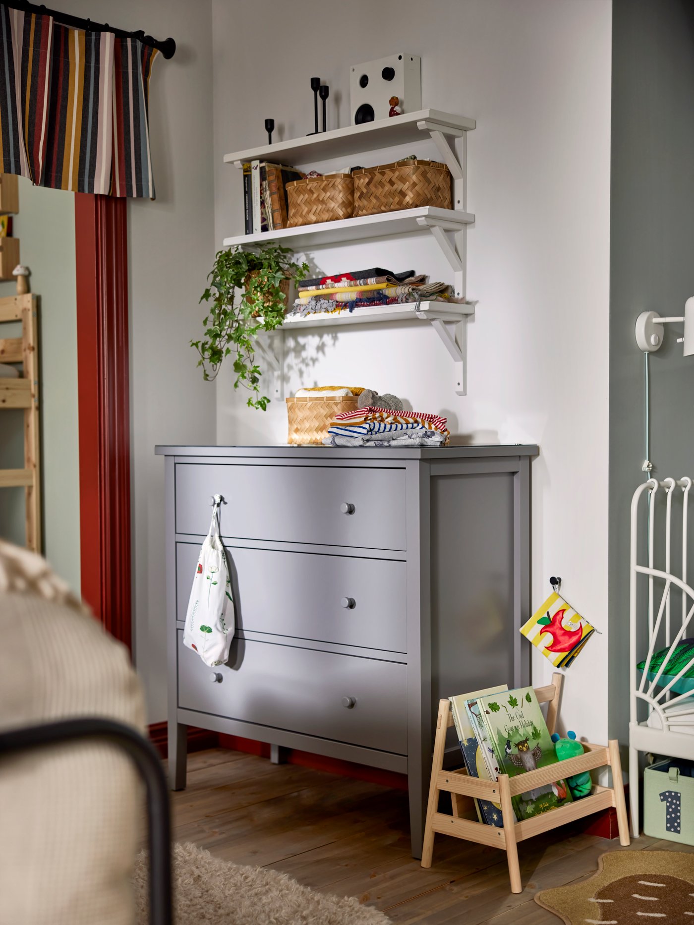 Grey chest of drawers with three wall mounted shelfs above, baskets, plants and decoration around.