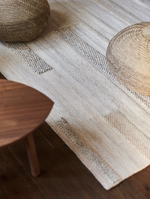 A beige BRÖNDEN low pile rug on a dark floor with two beige pouffes on it, and a STOCKHOLM coffee table beside it.