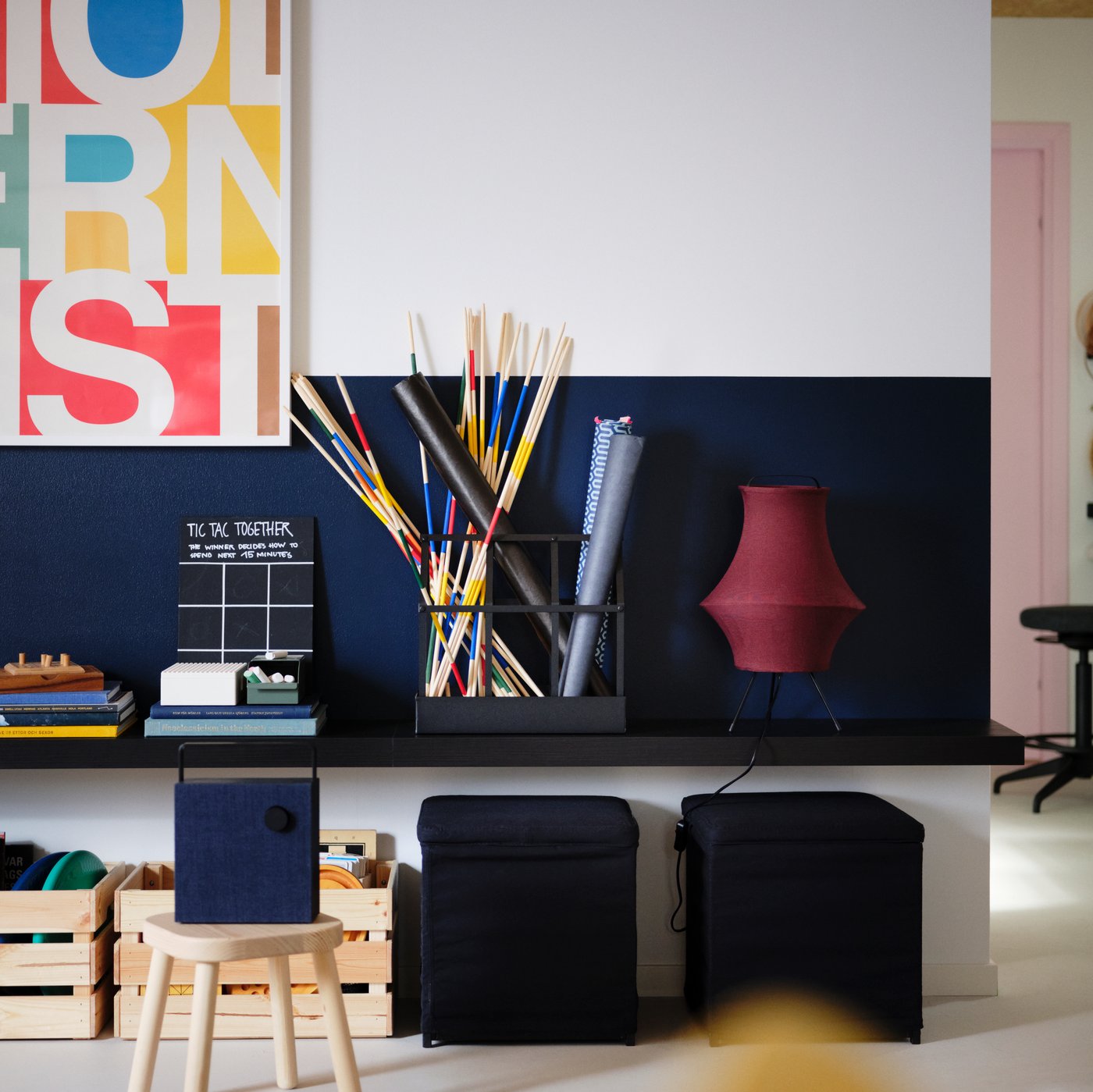 Dark red FYXNÄS table lamp on a black-brown LACK wall shelf, two black BOSNÄS footstools with storage and two pine boxes.