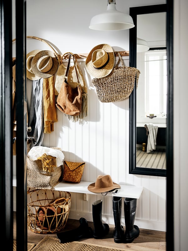 A hallway with natural-colored bags, hats and scarves on racks with six knobs and in baskets, beside a black TOFTBYN mirror.