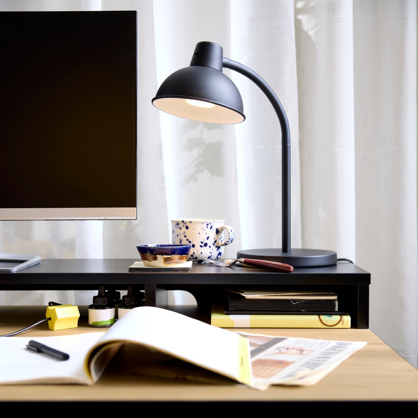 Detail horní skříňky psacího stolu MULLSJÖ s černou stolní lampou SKURUP a vedle ní vzorovaný modrý hrnek v kancelářském prostoru.