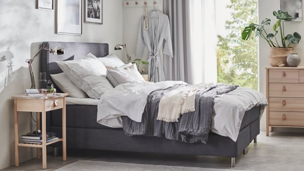Ein DUNVIK Boxspringbett in grau, daneben ein BJÖRKSNÄS Nachtkästchen und eine BJÖRKSNÄS Kommode.