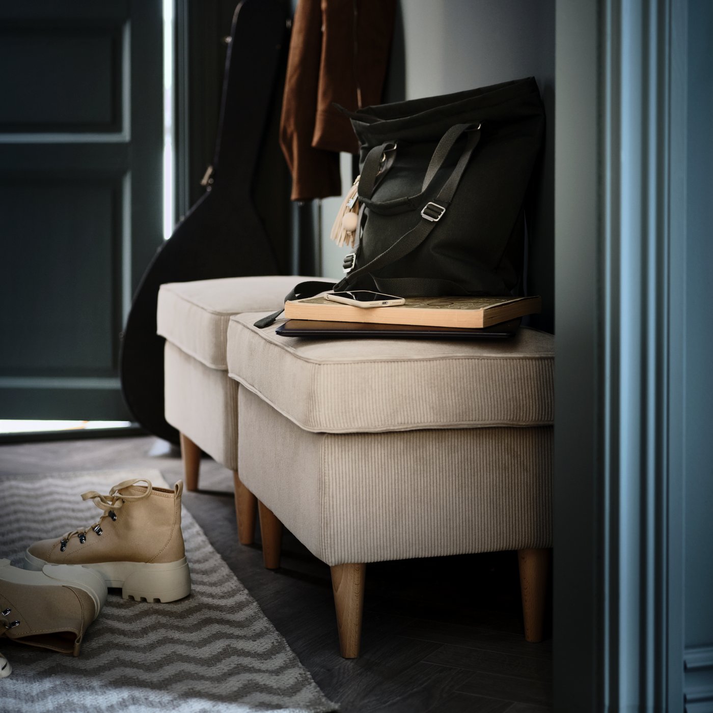 V predsieni sú pri stene dve béžové podnožky STRANDMON. Pod nimi je na koberci BREDEVAD s kľukatým vzorom položený pár topánok.