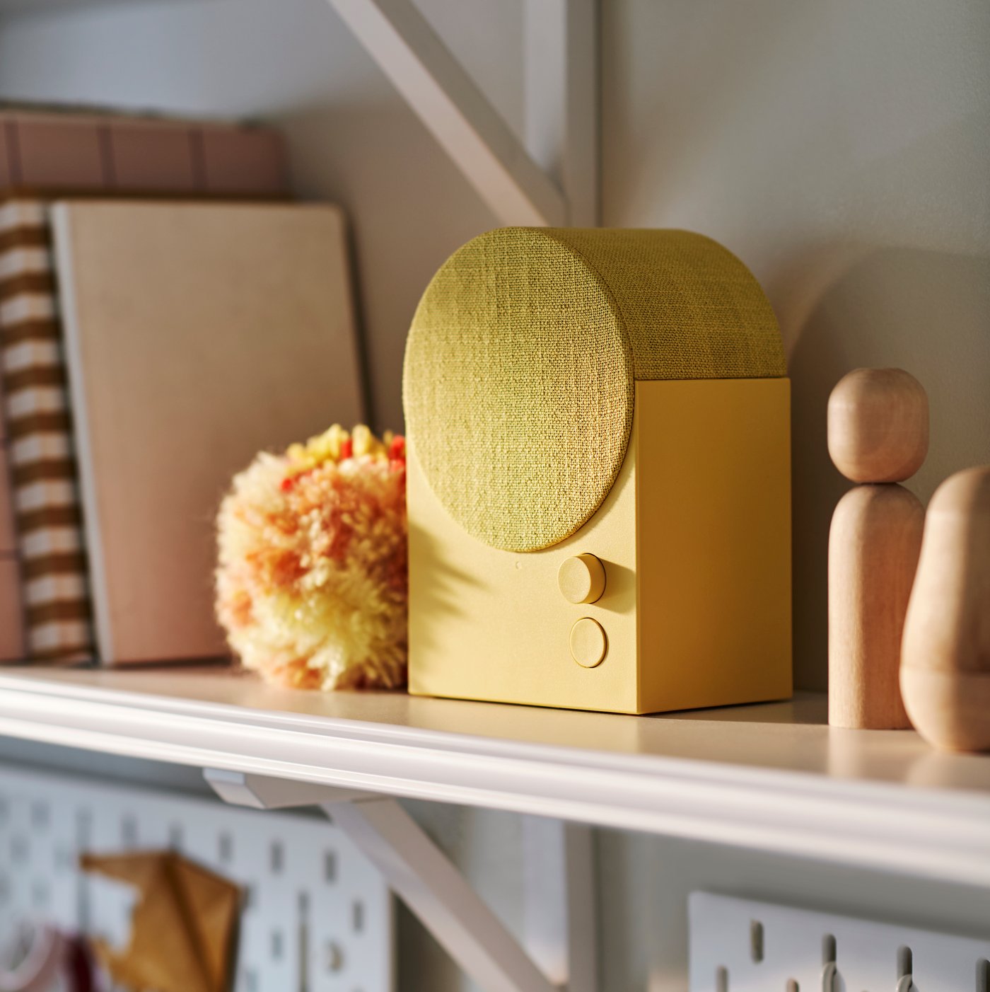 A yellow NATTBAD Bluetooth speaker with a rounded top sits on a shelf beside a plush pom-pom and minimalist wooden figures.