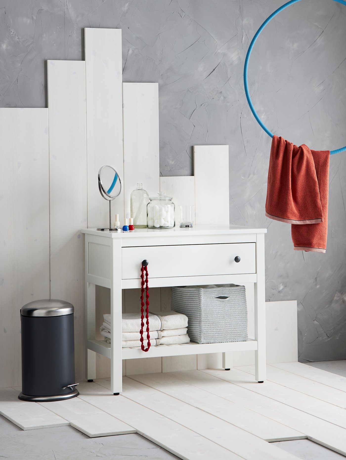 Baño con lavabo blanco abierto con cajón y estante, toallas de baño, toallas de mano, taza, tarro con tapa, espejo y cubo con pedal.