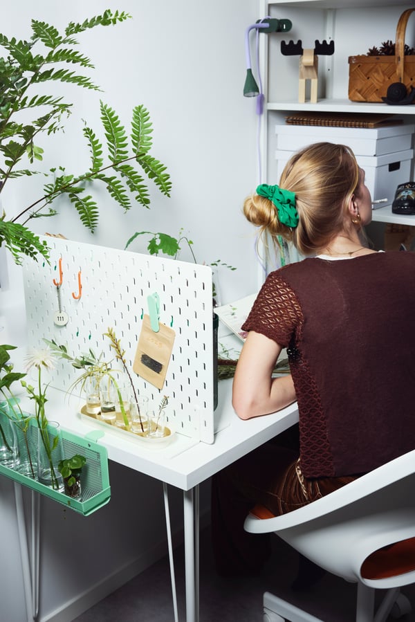 A person is seated at a desk in a plant-filled workspace, surrounded by glass jars, a white SKÅDIS pegboard with hanging items, and storage boxes in the background.