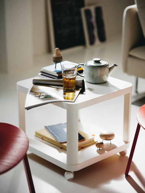 O măsuță de cafea  albă, pătrată, cu rotile, care conține un ceainic GLADELIG, un pahar POKAL cu lichid maro și reviste.
