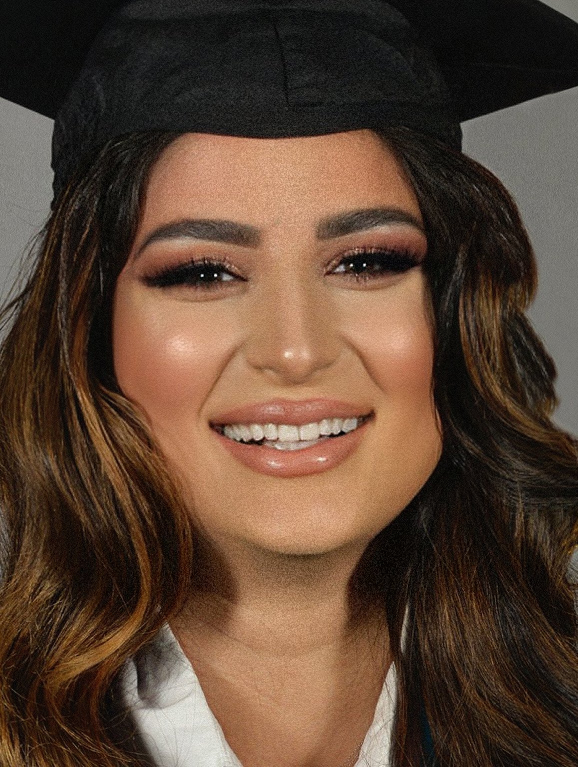 Portrait of Iraqi refugee Aya Abdullah smiling, wearing a black and yellow graduate cap against a gray background.