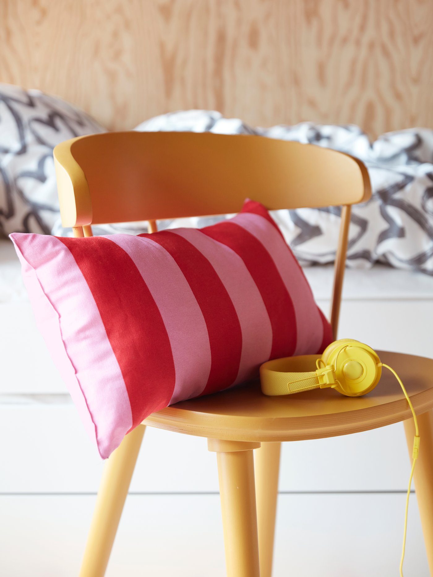Ein gelber Stuhl, Kopfhörer und ein rosa-rot gestreiftes SARAKAJSA Kissen vor einem Bett mit weiß-grauer Bettwäsche.