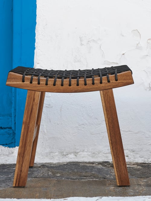 A white wall and a light brown, stained STACKHOLMEN stool with a black, braided rope seat that's slightly curved upward.