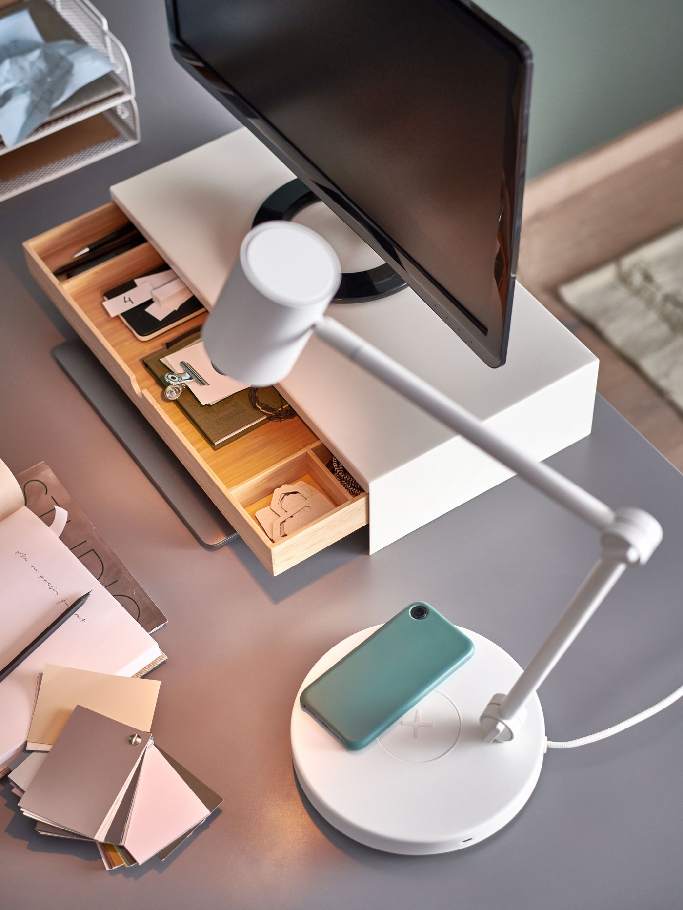 A smart phone lays on the built-in charger of a white NYMÅNE work lamp with wiress charging that is shining light on a desk.