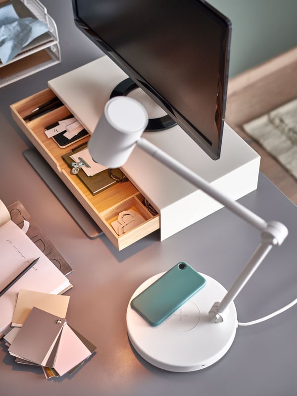 A smart phone lays on the built-in charger of a white NYMÅNE work lamp with wiress charging that is shining light on a desk.