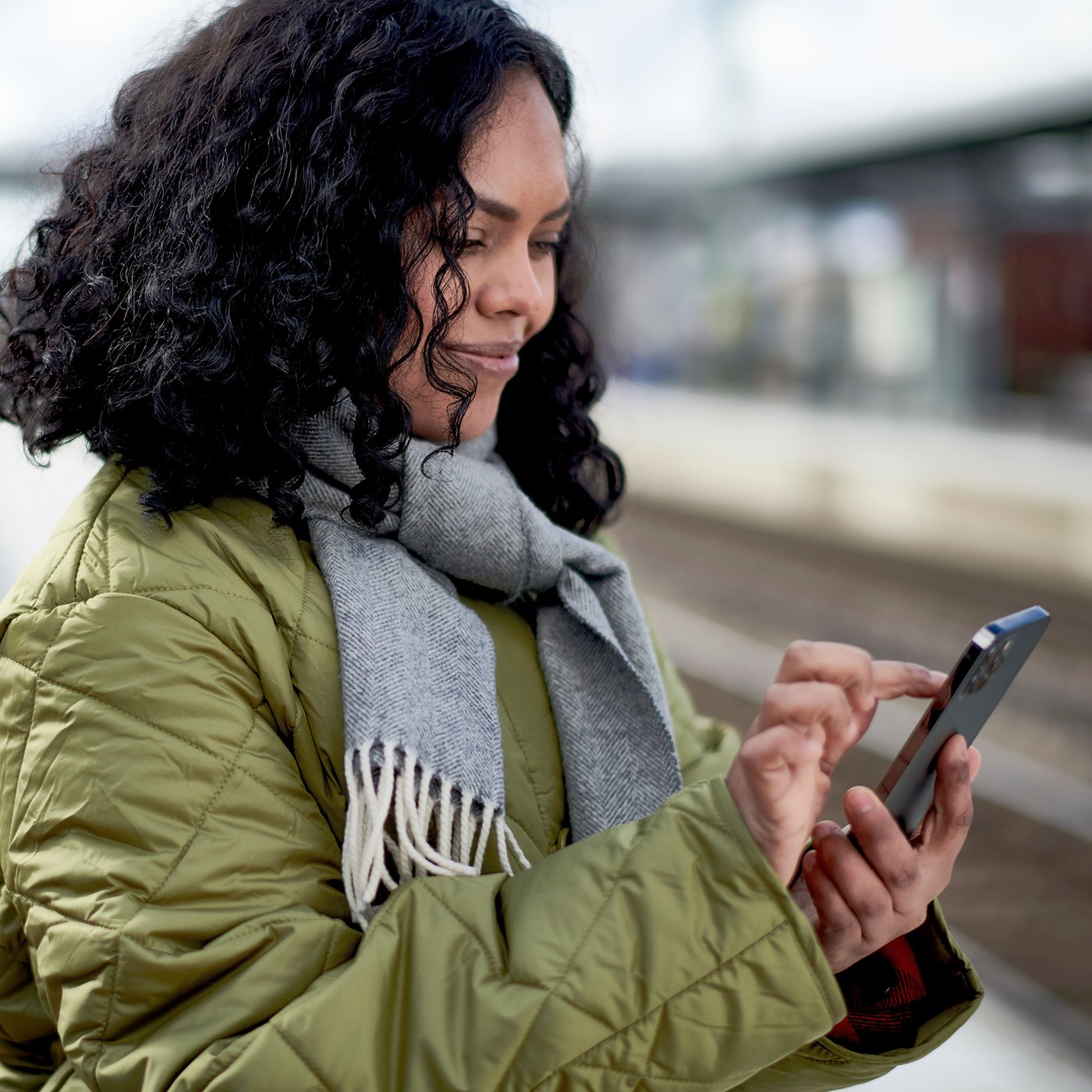 A woman with curly hair in a green jacket and gray scarf is scrolling on her phone while waiting outside at a train platform.