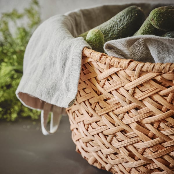 Um cesto TJILLEVIPS em rota, com toalhas de rosto em branco e pepinos no chão à frente de uma planta verde.