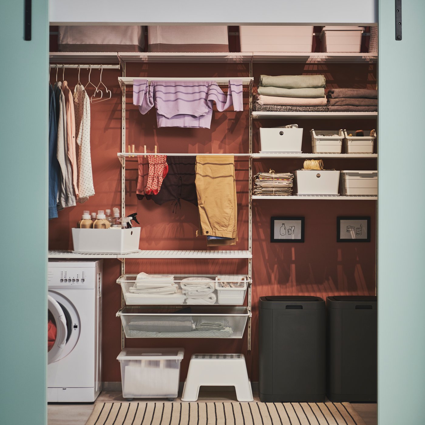 Nástěnná policová sestava BOAXEL kolem pračky. Na sestavě jsou poukládané kusy sušícího se oblečení a různé koše a krabičky.