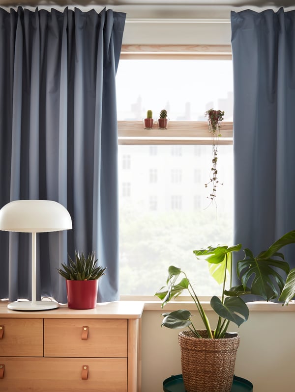 Blue BENGTA curtains half-drawn across a window behind a BJÖRKSNÄS chest of drawers with plants and a white lamp on top.